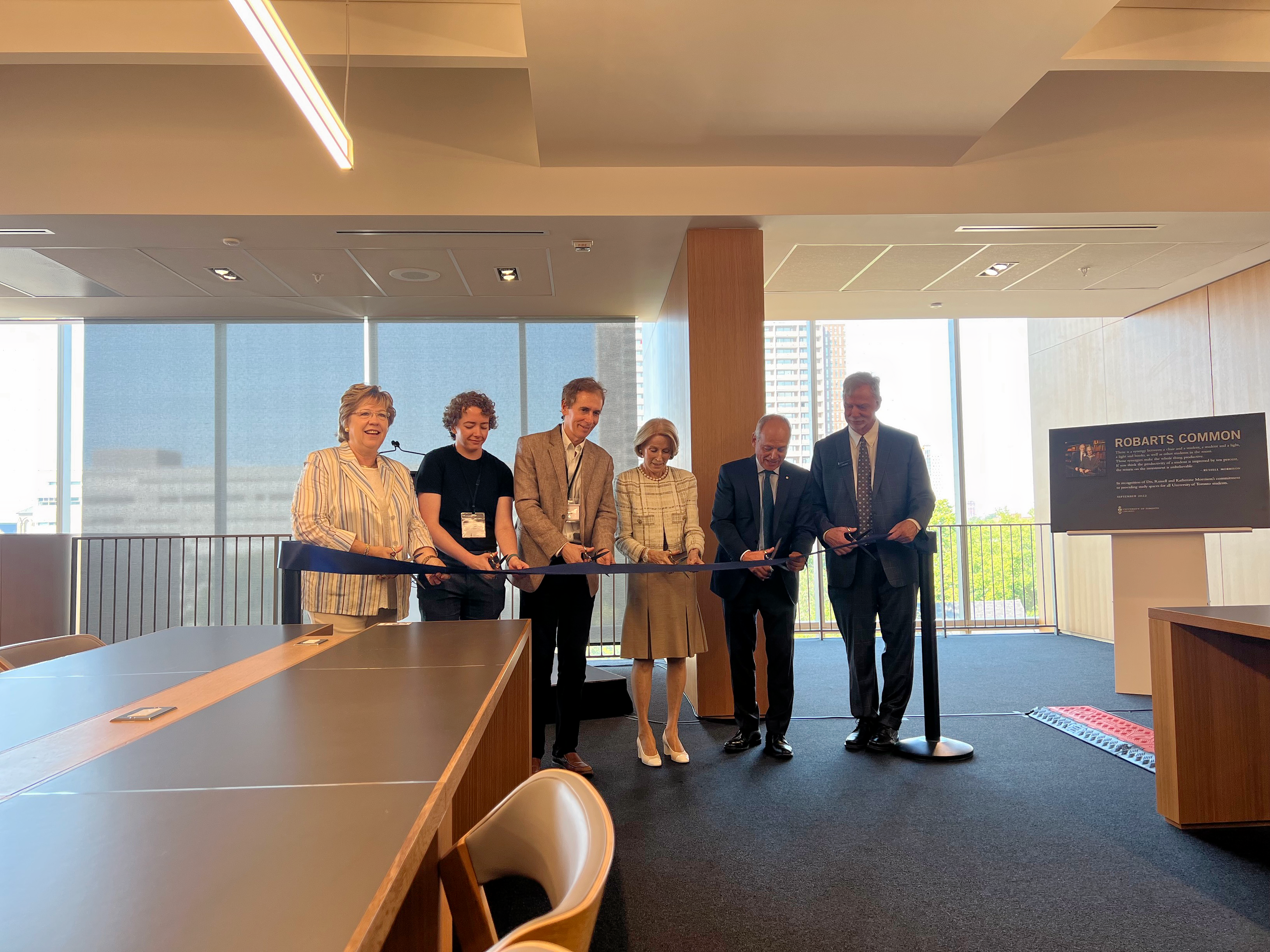 Two women and four men stand in a line. They each are holding a pair of scissors poised to cut a long ribbon which is in front of them, tied to a short pole on each end. They are dressed in business casual clothing and they are all smiling. They are indoors; behind them are floor to ceiling windows and a single wooden wall divider. They are standing on blue carpet, and one long table with a chair is in sight.