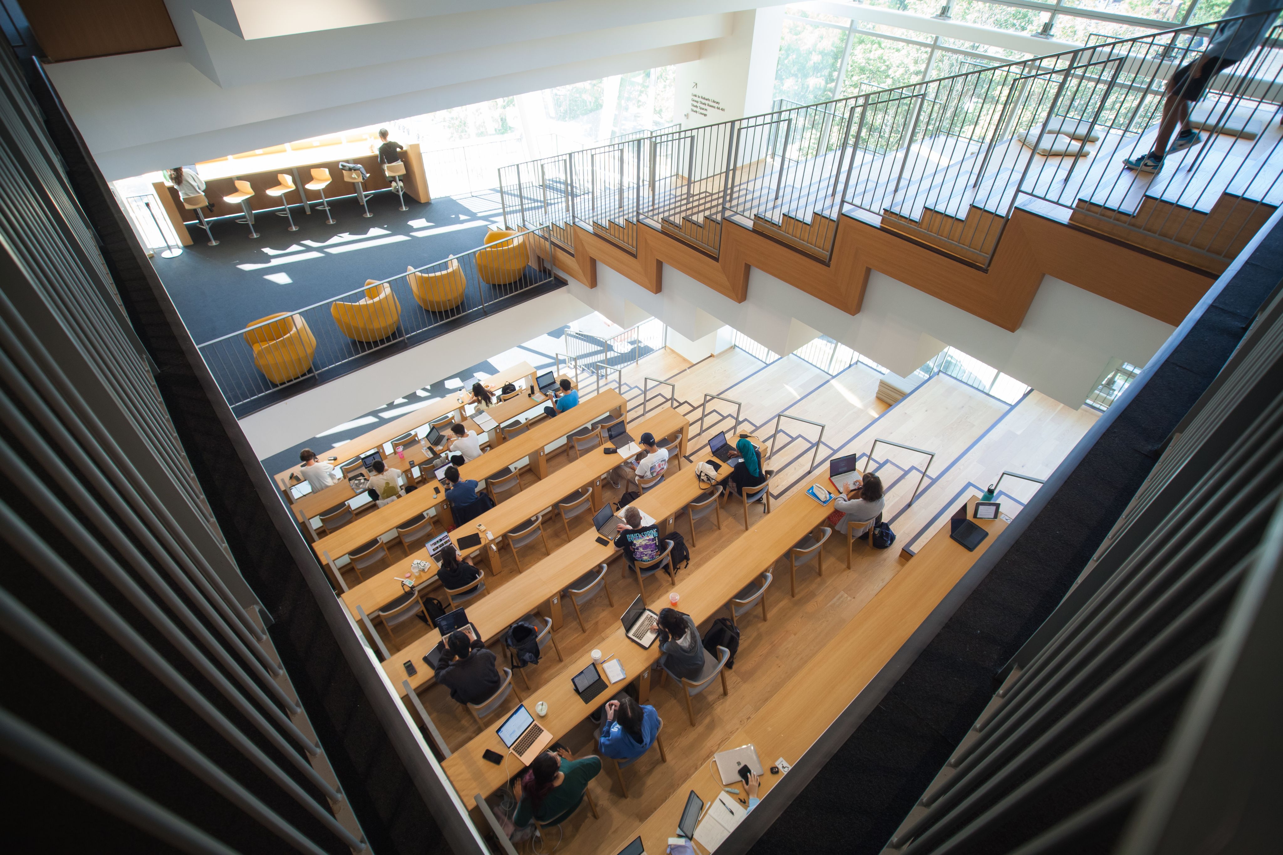 Architectural photo of the second floor lecture style study desks taking from the top of the third floor. there are dozens of students sitting and studying with laptops, notebooks or text books in front of them. There are some students walking down the stairs and two students situated at the front window overseeing the Plaza in front of Robarts Common. The space is bright with sun shining from all the windows