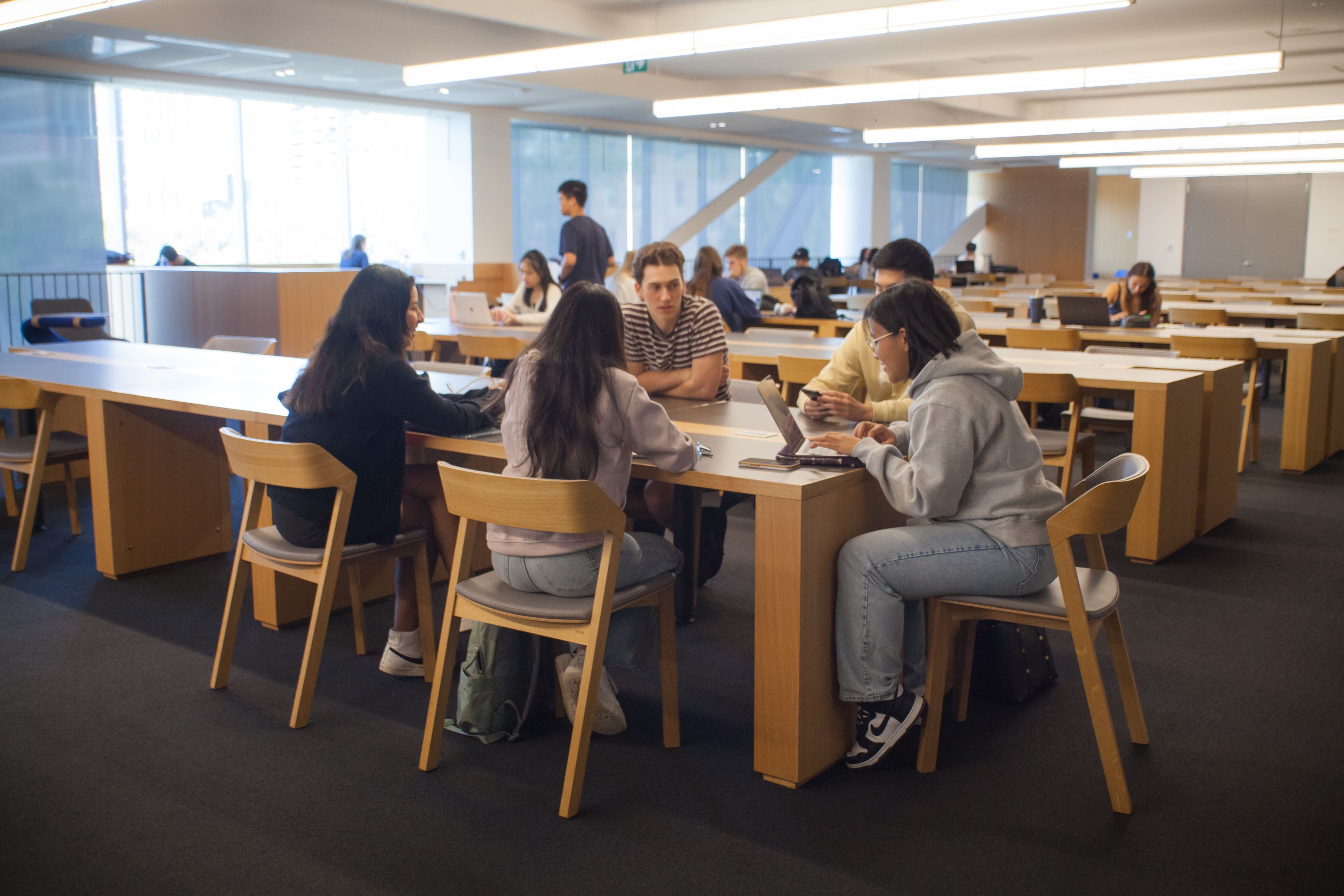 Image of five students sitting at a tabletwo are typing in a laptop where others are engaged in conversation. the students are situated in the middle of Robarts Common there are surrounded by long tabled with students studying in the background. The atmosphere is bright and the sun is shining outdoors