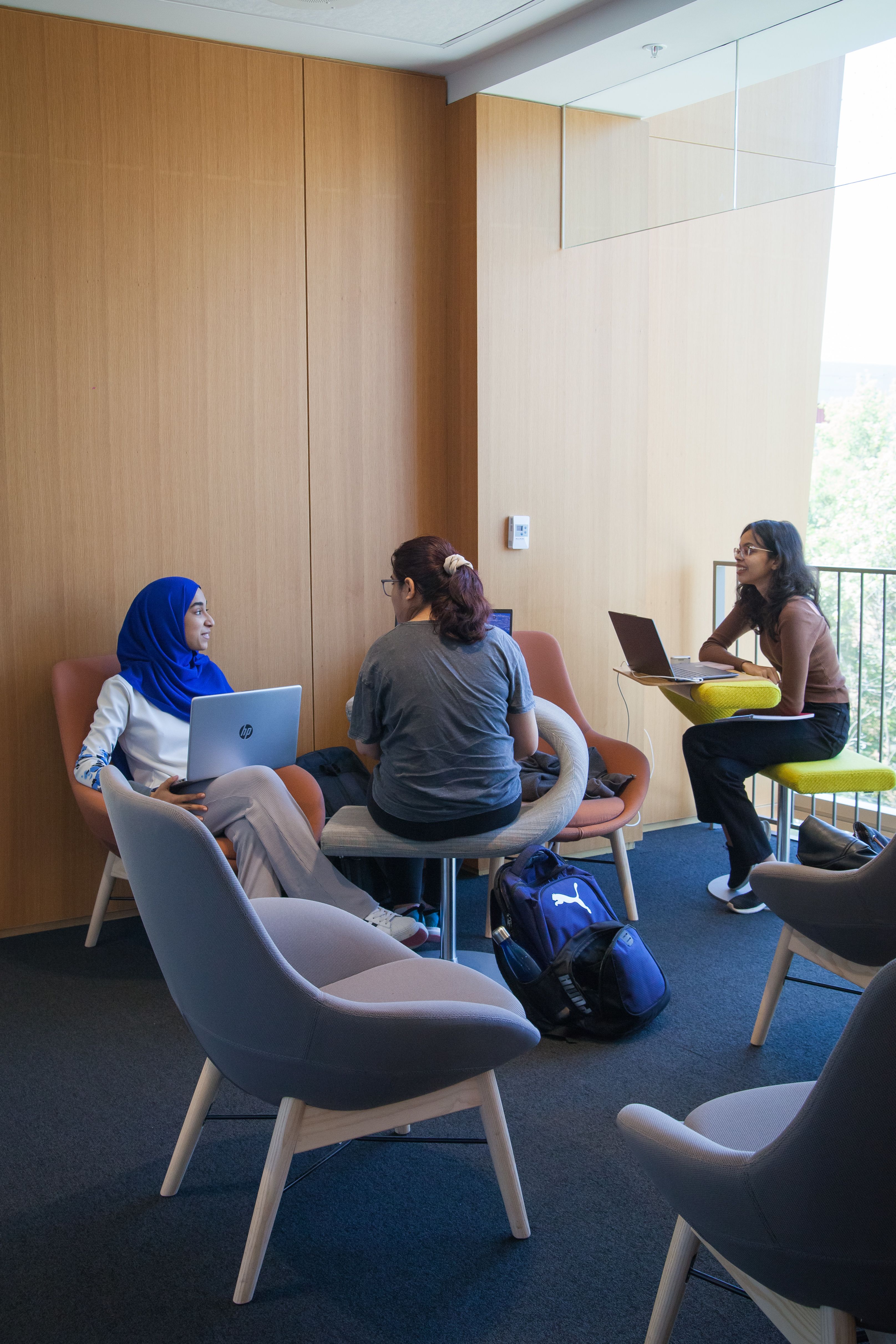 three students all with laptops laughing and chatting in the corner wings of the Robarts Common study room. Two students are facing each other and laughing while one student has her back to the camera.