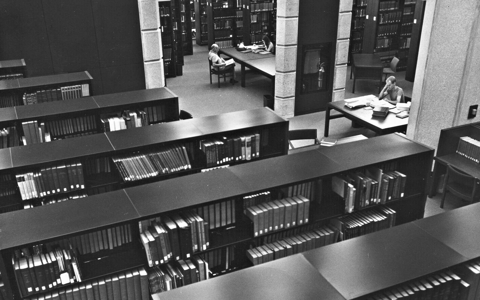Black and white image of two students sitting on old large wooden desks with stacks of books in the background in a reading room at Robarts Library
